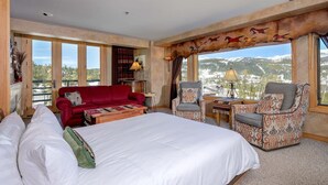 A hotel room with a large bed, a red sofa, two armchairs, a small table, and a view of mountains. - Madison Vista-Big Sky Shoshone Ski in/Out Condo (Big Sky)