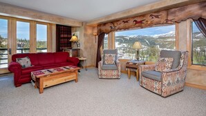 A spacious room with a red sofa, two patterned armchairs, a wooden coffee table, and a view of mountains through large windows. - Madison Vista-Big Sky Shoshone Ski in/Out Condo (Big Sky)