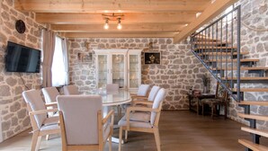 A spacious room with a stone wall, a round dining table with chairs, a television mounted on the wall, and a staircase with a metal railing. - Awesome home in Imotski (Imotski)