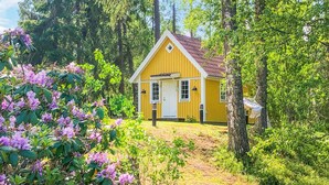 Une cabane jaune avec une porte blanche, entourée d’une végétation luxuriante et d’arbres.