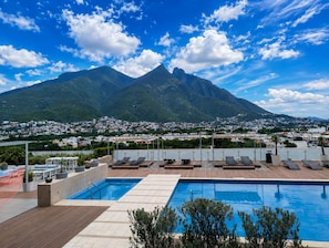 A rooftop pool with lounge chairs, a view of a mountain, and a cityscape in the background.