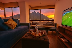 A living room with a sofa, ottoman, and a coffee table with a bottle and glasses. A television is on the right, and a window offers a view of a mountain and city lights at sunset.