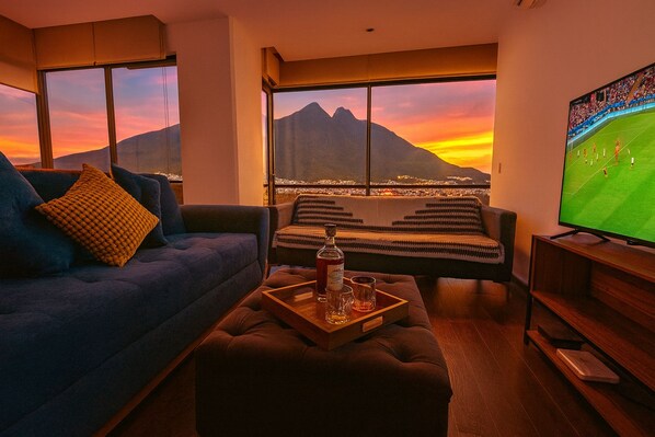 A living room with a sofa, ottoman, and a coffee table with a bottle and glasses. A television is on the right, and a window offers a view of a mountain and city lights at sunset.