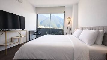 A modern hotel room with a large bed, a flat-screen TV, a desk, and a view of mountains through large windows.