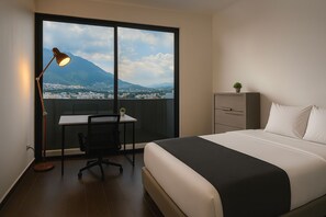 Ett hotellrum med en säng, ett skrivbord, en stol, en lampa, en byrå och utsikt över ett berg och en stadsbild genom ett stort fönster.