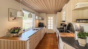 Une cuisine avec des meubles de rangement en bois, un sol carrelé, des luminaires suspendus et une vue par la fenêtre.