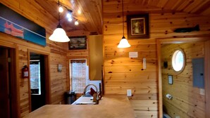Interior de una cabaña de madera con una isla central, iluminación colgante y un ventanal redondo.