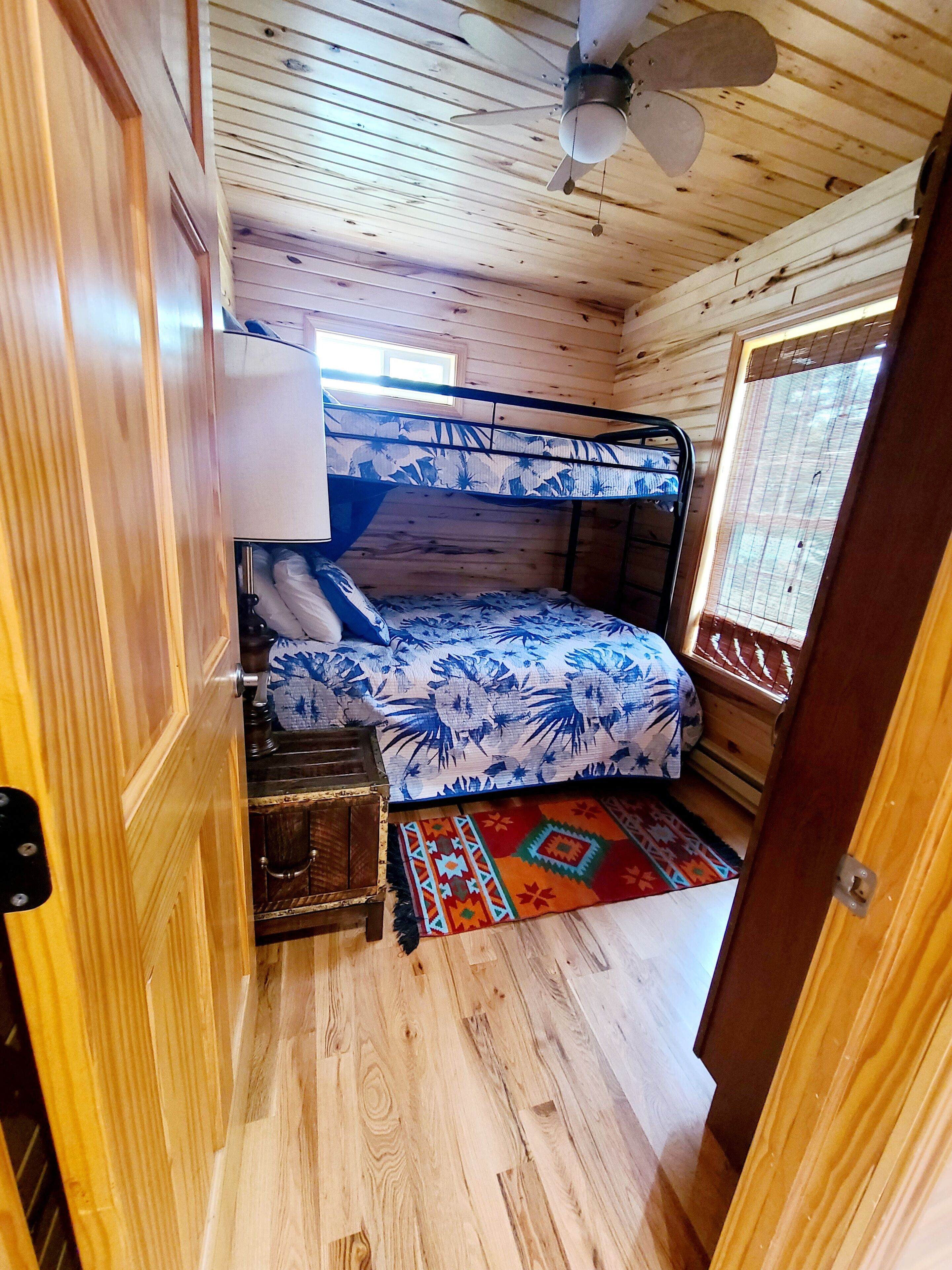Una habitación de cabaña de madera con literas, un ventilador de techo, una ventana con persianas y un pequeño cofre de madera.