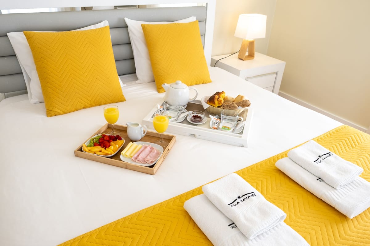Un lit bien fait, avec un plateau contenant divers éléments pour le petit-déjeuner, notamment des fruits, des viennoiseries et une boisson.
