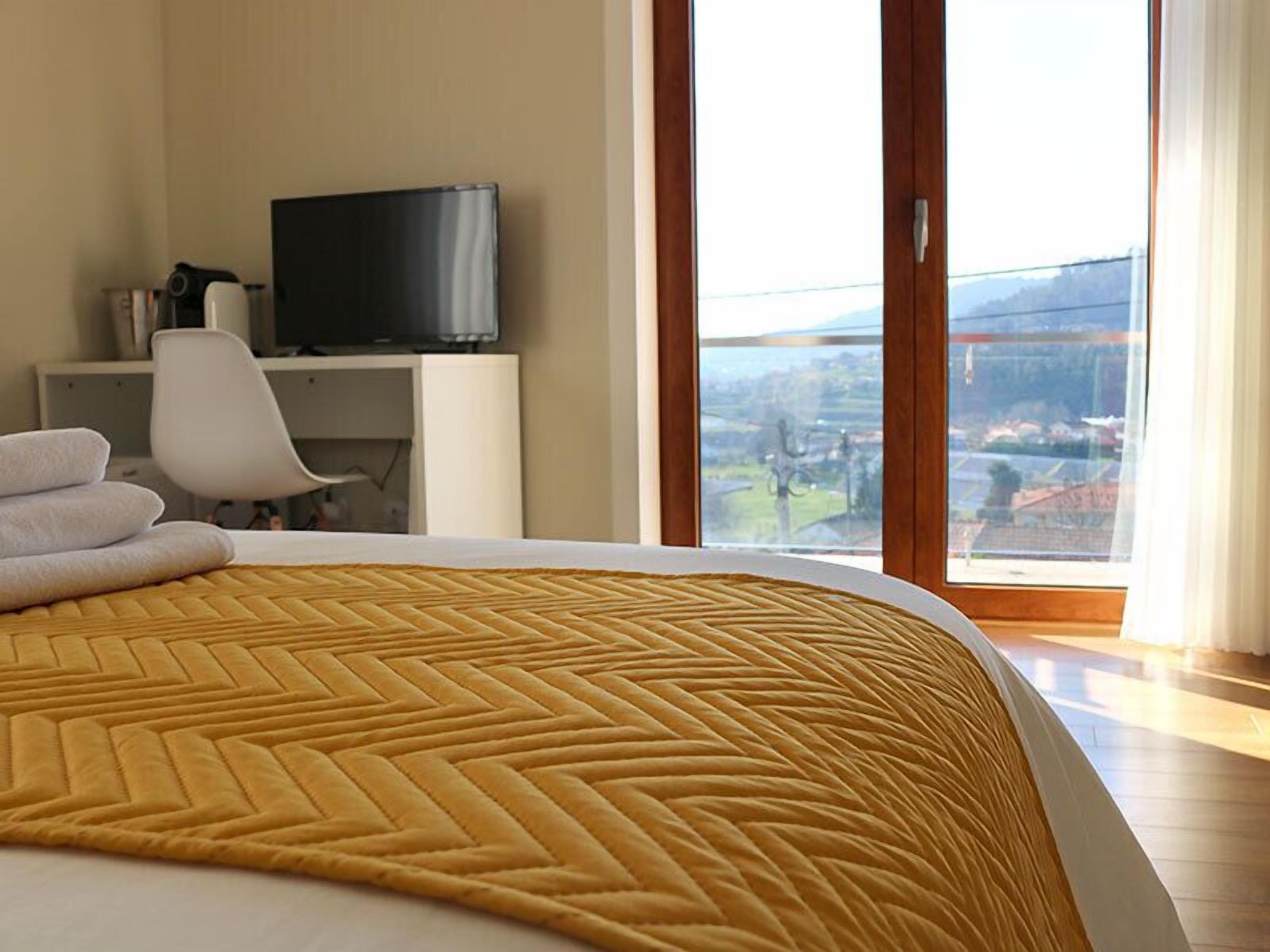 A hotel room with a bed, a television on a desk, and a view of a cityscape through large windows.