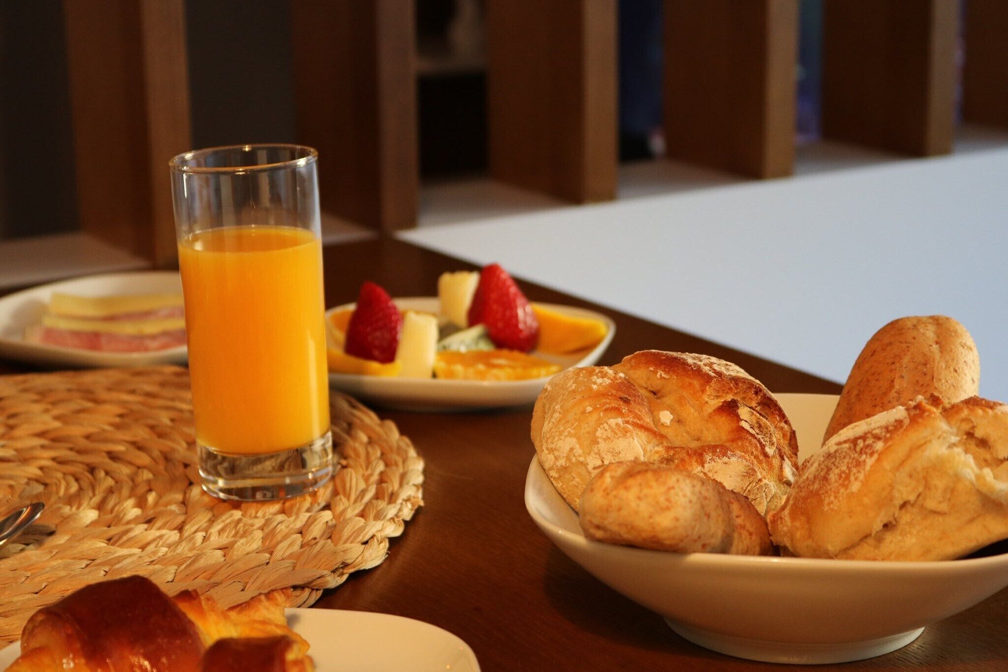 A breakfast setting with a glass of orange juice, a plate of strawberries, and a bowl of croissants on a woven placemat.