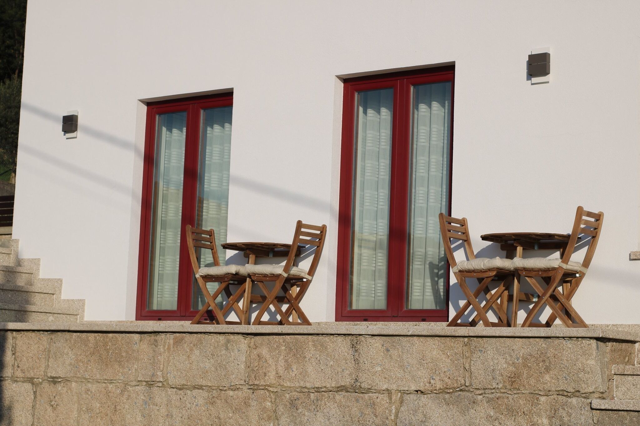 A building with two windows, each with a red frame and a small air conditioning unit on the wall. There are two wooden chairs and a small round table in front of each window.