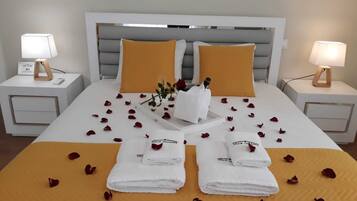 Ein ordentlich gemachtes Bett mit weiß-gelber Farbgebung, zwei weiße Handtücher, Rosenblätter und eine kleine Vase mit Blumen.