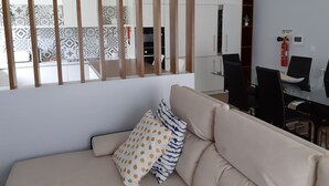 A modern living room with a beige sofa, patterned cushions, and a view of the kitchen and dining area through a wooden partition. - Villa Tavares Guest Houses (Arouca)