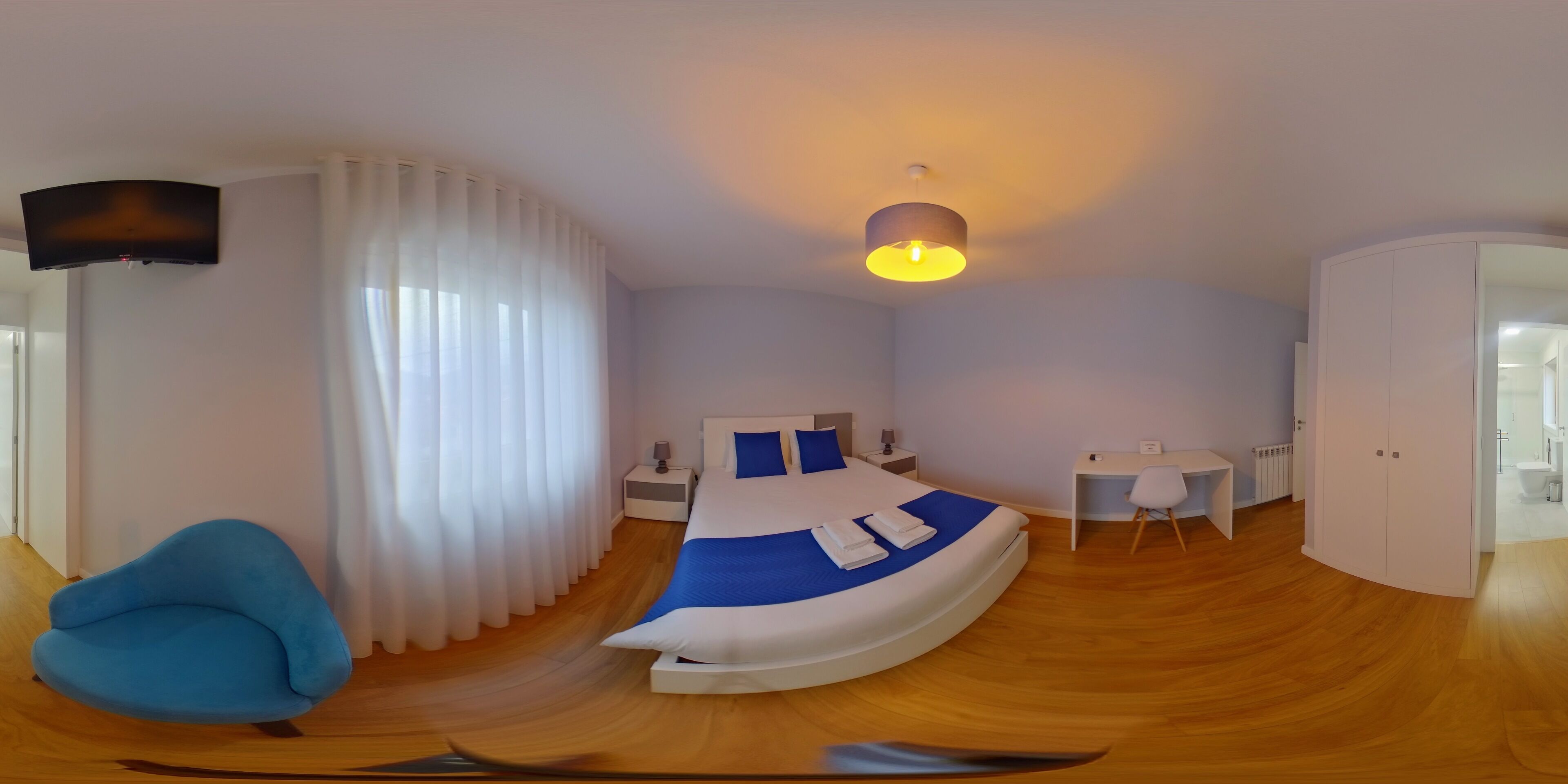 A modern hotel room with a curved bed, a desk, a chair, a TV, and a bathroom visible through an open door.