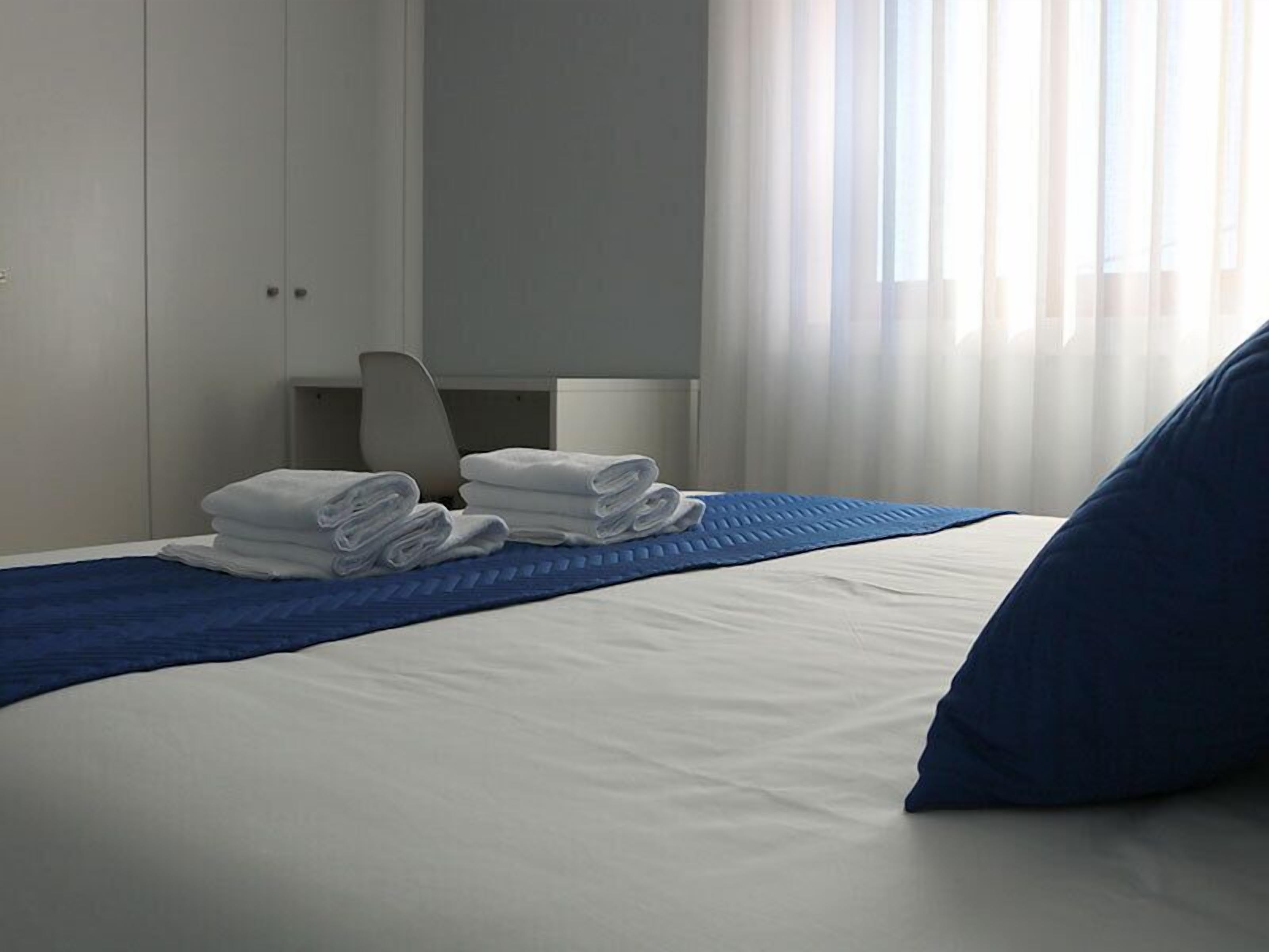 Een hotelkamer met een net opgemaakt bed, een blauw sierkussen en een stapje gevouwen handdoeken op het bed.
