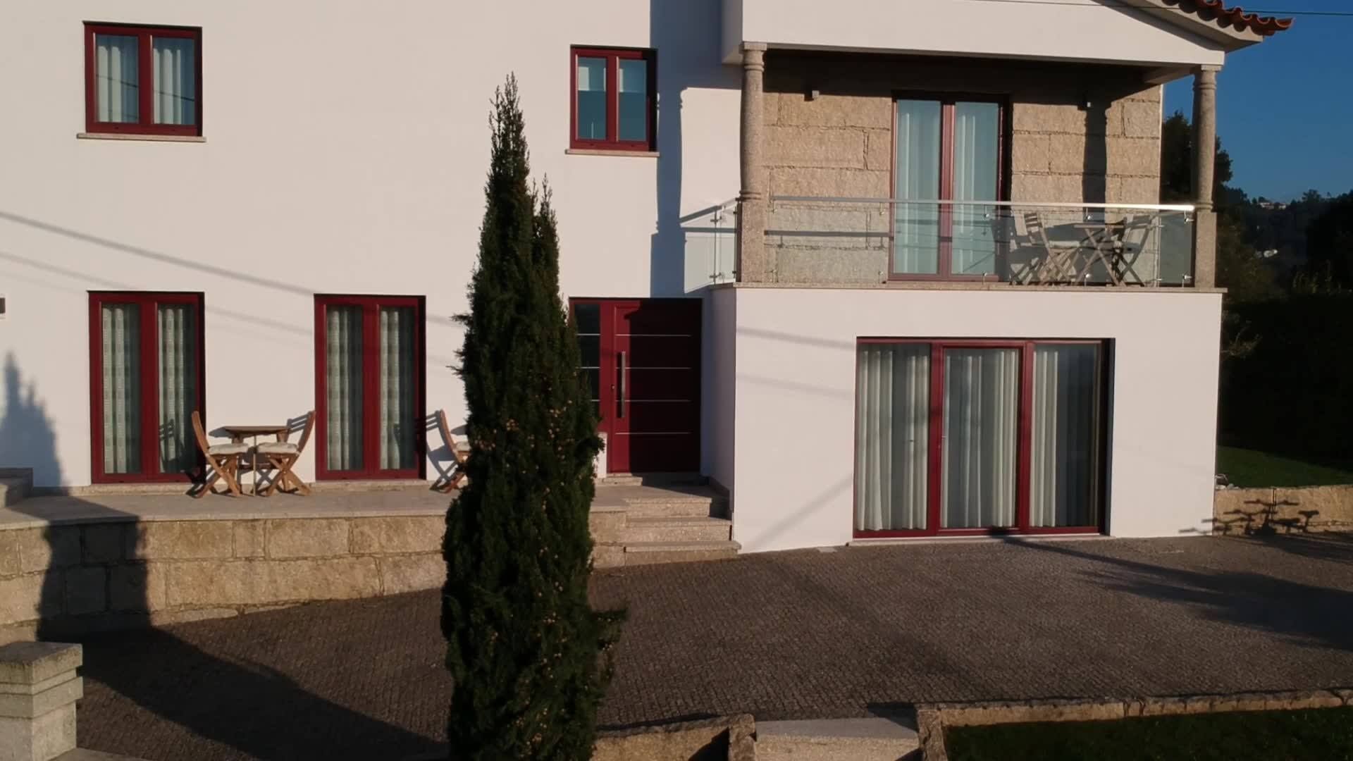 A modern two-story house with a balcony, a patio with outdoor furniture, and a tall evergreen tree in the foreground.