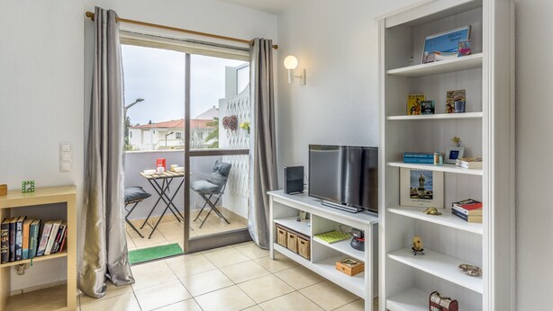 Ein modernes Wohnzimmer mit Schiebeglasfenster zum Balkon, einem Flachbildfernseher, einem Bücherregal und einem kleinen Tisch mit einem Stuhl.