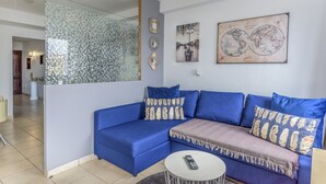 Ein Wohnzimmer mit einem blauen Sofa, einem runden Couchtisch und einem Spiegel. Dekorative Kissen und eine Weltkarte sind an der Wand zu sehen.