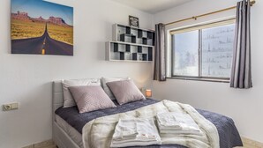 Ein Schlafzimmer mit Bett, Kissen, Decke, einem Regal mit Büchern, einem Gemälde und einem Fenster mit Vorhängen.