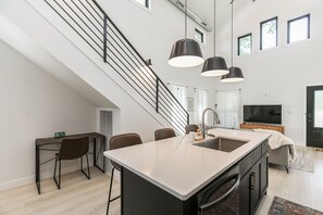 A modern kitchen with a central island, a dining area with chairs, a staircase, and a large window providing natural light.