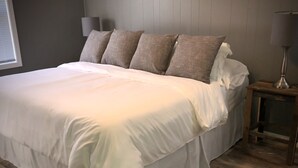 A neatly made bed with white bedding and several pillows, a wooden floor, a small bedside table with a lamp, and a wall-mounted lamp.