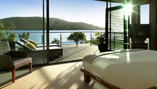 Un dormitorio moderno con una cama grande, televisión y balcón con vistas a un lago y colinas.