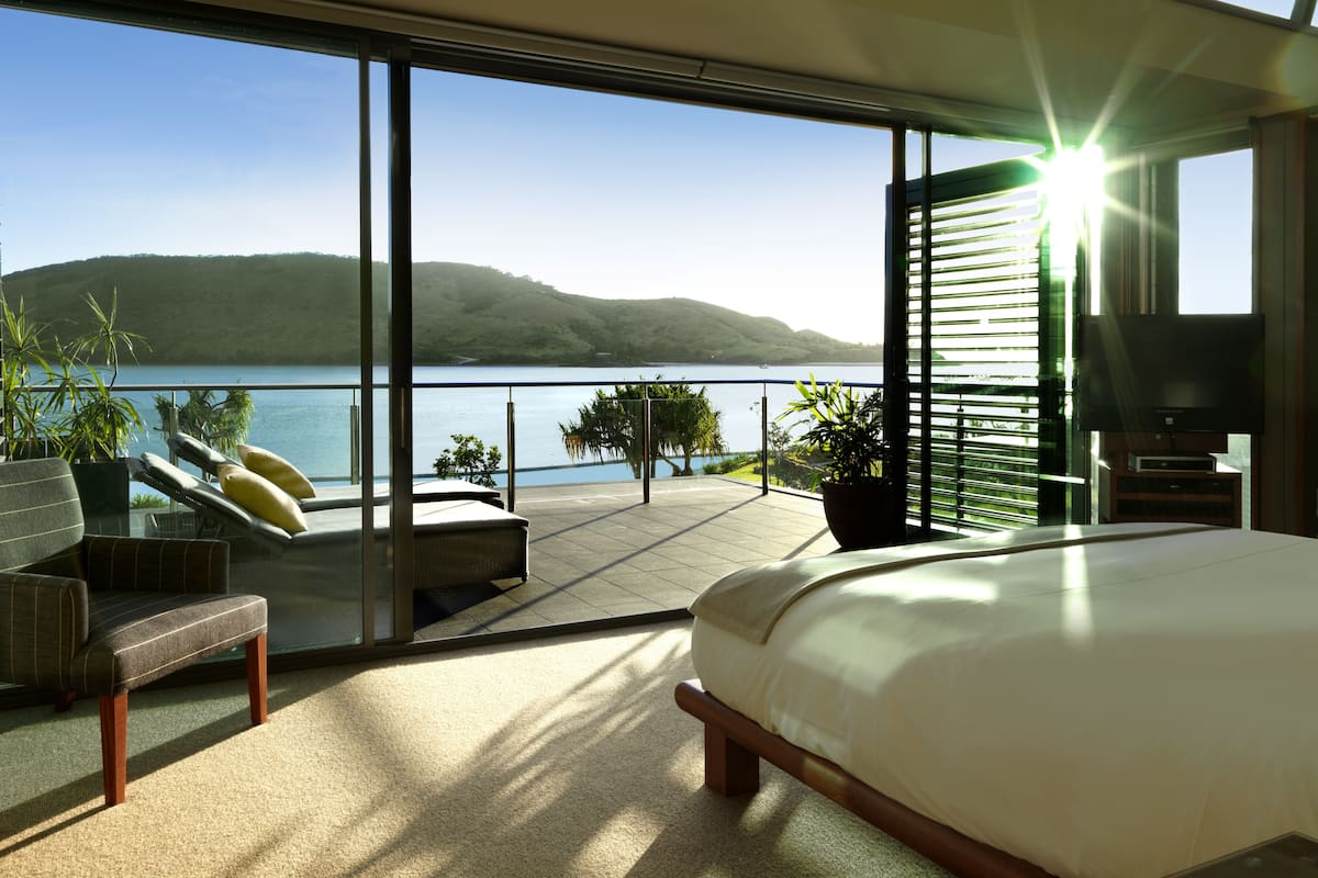 A modern bedroom with a large bed, a TV, and a balcony with a view of a lake and hills.