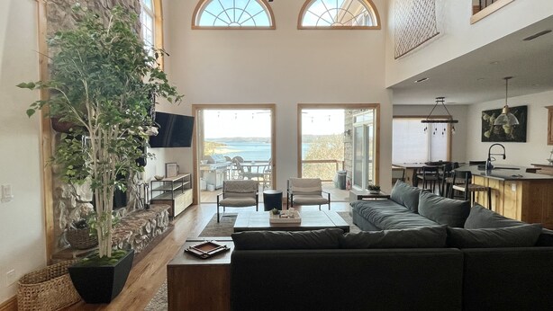 A spacious living area with a large sofa, a dining table, and a kitchen area. There is a large window with a view of the outdoors. - Camp Roberts-Lakefront-8 Bedrooms-Sleeps 19-Big; Lake View-Community Pool! (Garfield)