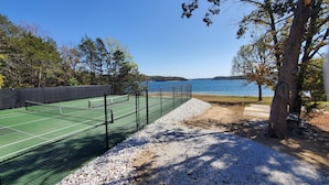 A tennis court with a net, surrounded by gravel and trees, overlooking a body of water. - Camp Roberts-Lakefront-8 Bedrooms-Sleeps 19-Big; Lake View-Community Pool! (Garfield)