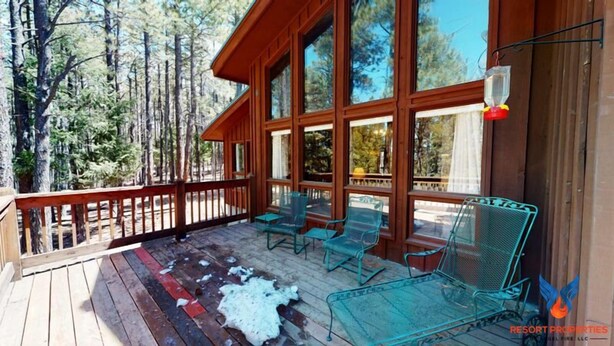 A wooden deck with glass chairs and a table, surrounded by large windows and a view of trees. - Close to Country Club | Fireplace | Single Level | Happy Pines (Angel Fire)