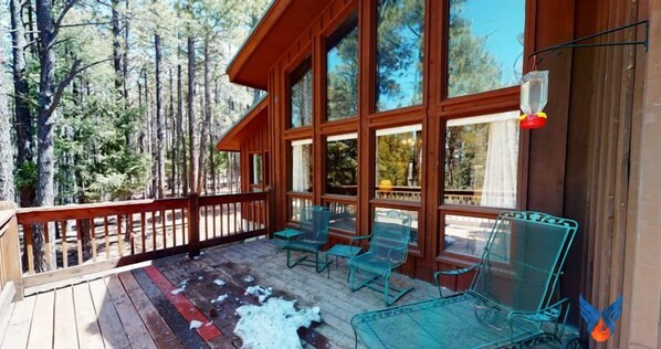 A wooden deck with glass chairs and a table, surrounded by large windows and a view of trees.