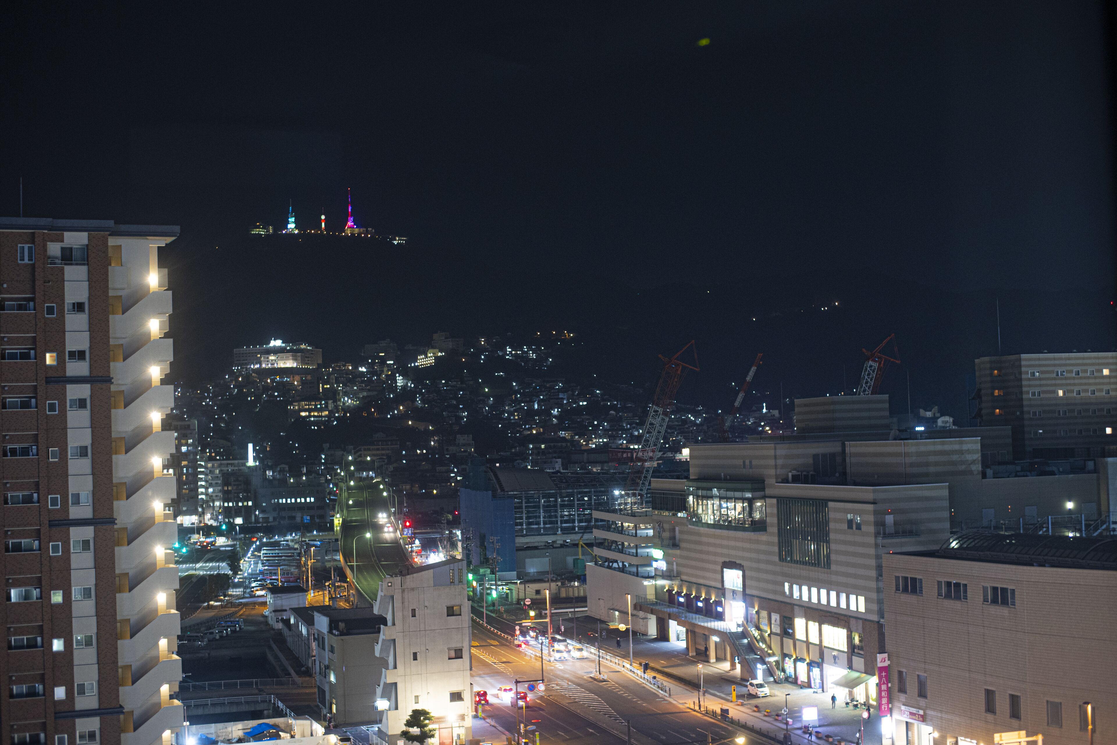 A nighttime cityscape with illuminated buildings and a distant view of a hill with colorful lights.