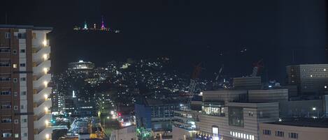 Une vue nocturne d’une ville, avec des bâtiments illuminés et, au loin, une colline parée de lumières colorées.