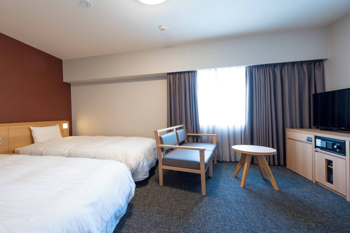 A hotel room with two beds, a desk, a chair, a television, and a window with curtains.