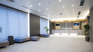 A modern reception area with a reception desk, seating, and a large window with blinds. - Dormy Inn Nagasakiekimae Hot Springs (Nagasaki)