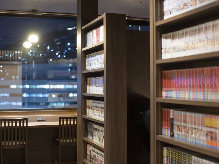 A library interior with bookshelves, a window with a view of a cityscape, and two wooden chairs.
