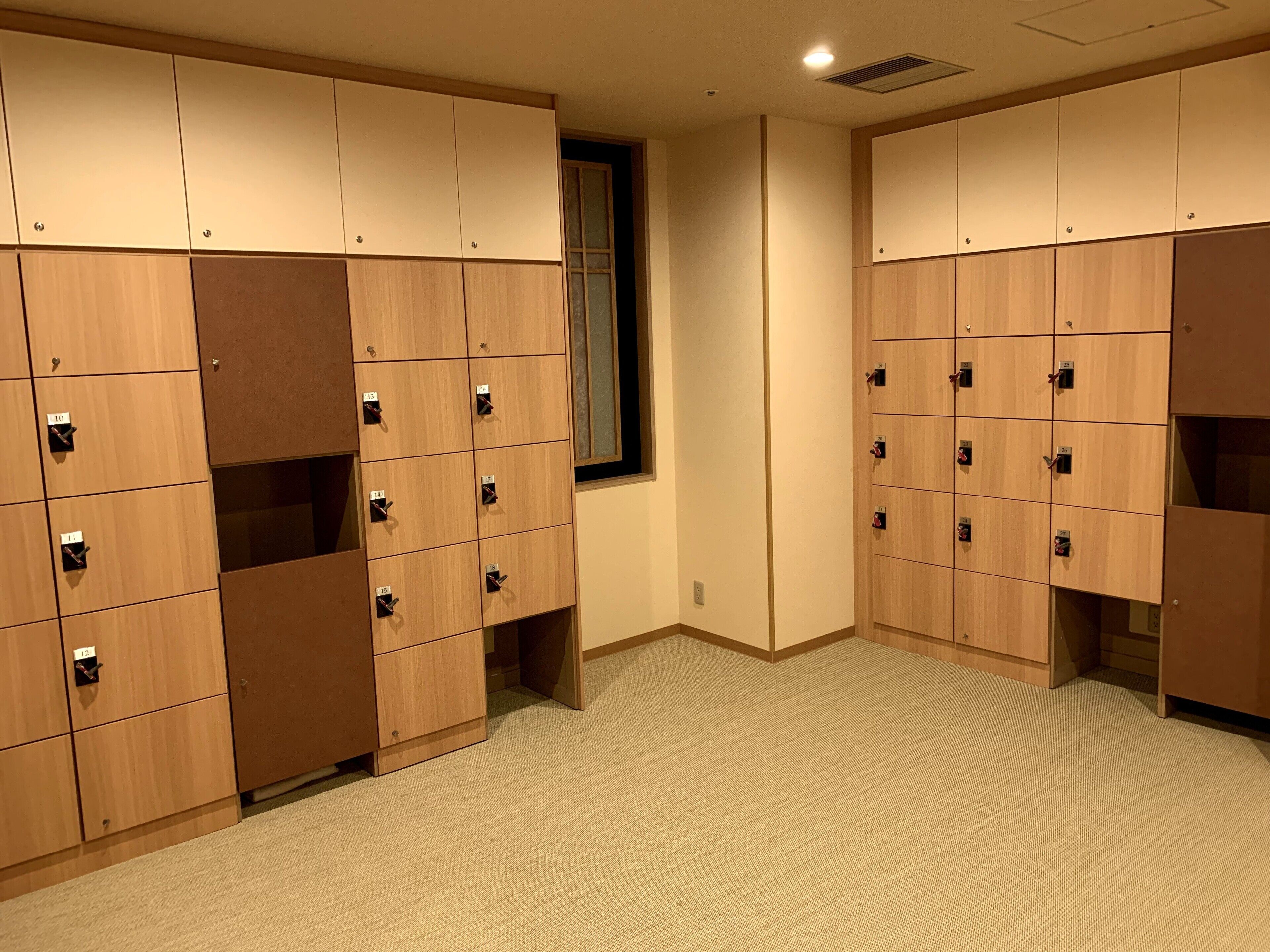 A row of wooden lockers with numbered compartments, carpeted floor, and a window with blinds.