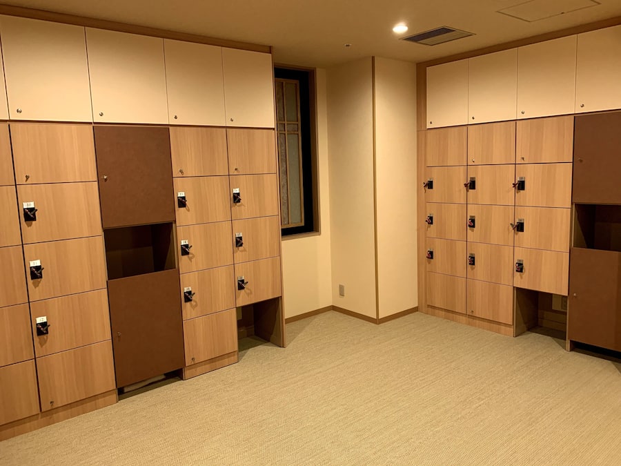 A row of wooden lockers with numbered compartments, carpeted floor, and a window with blinds.