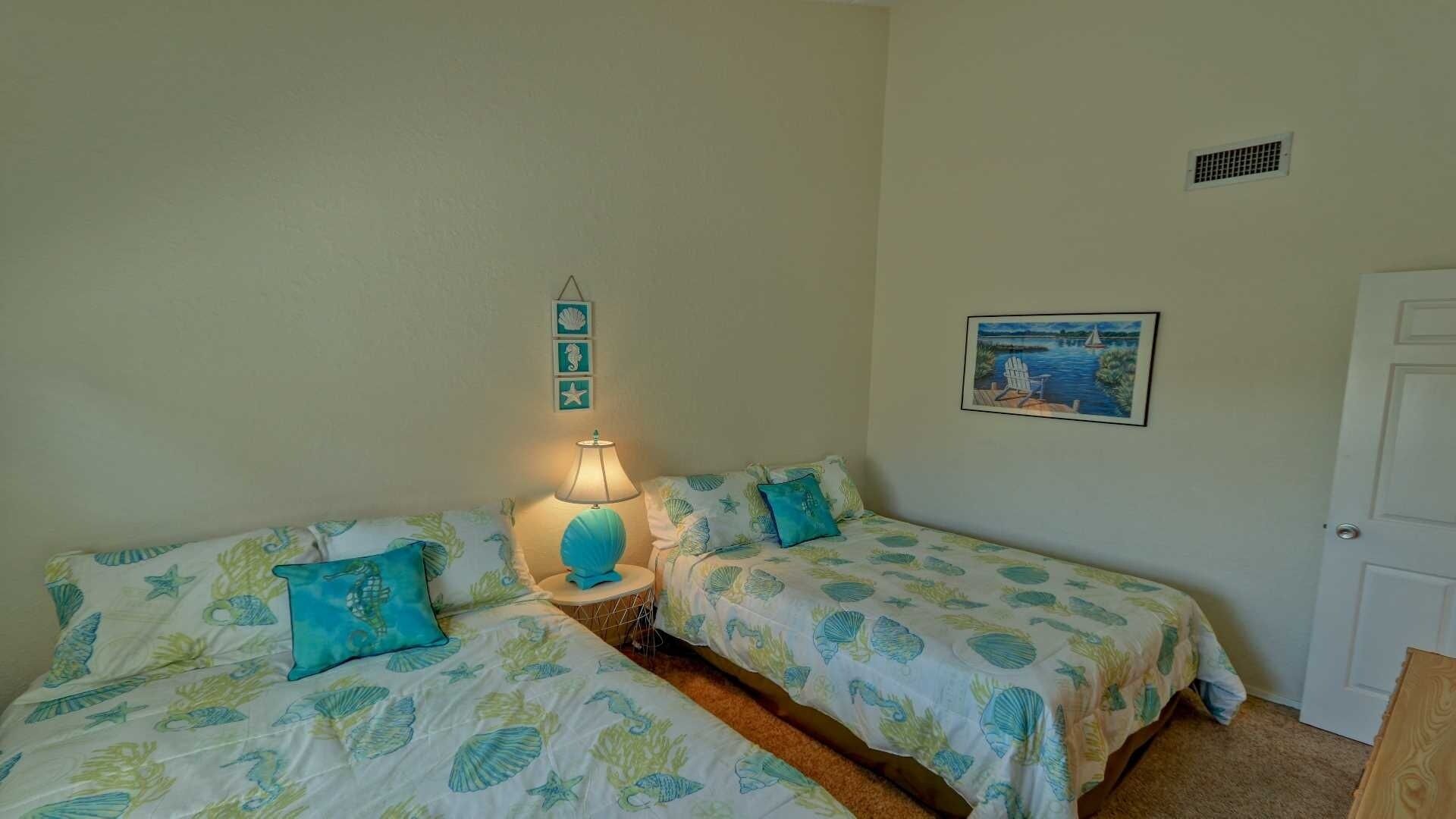 A room with two beds, each with matching blue and green bedding, a bedside lamp, and a small wall decoration.