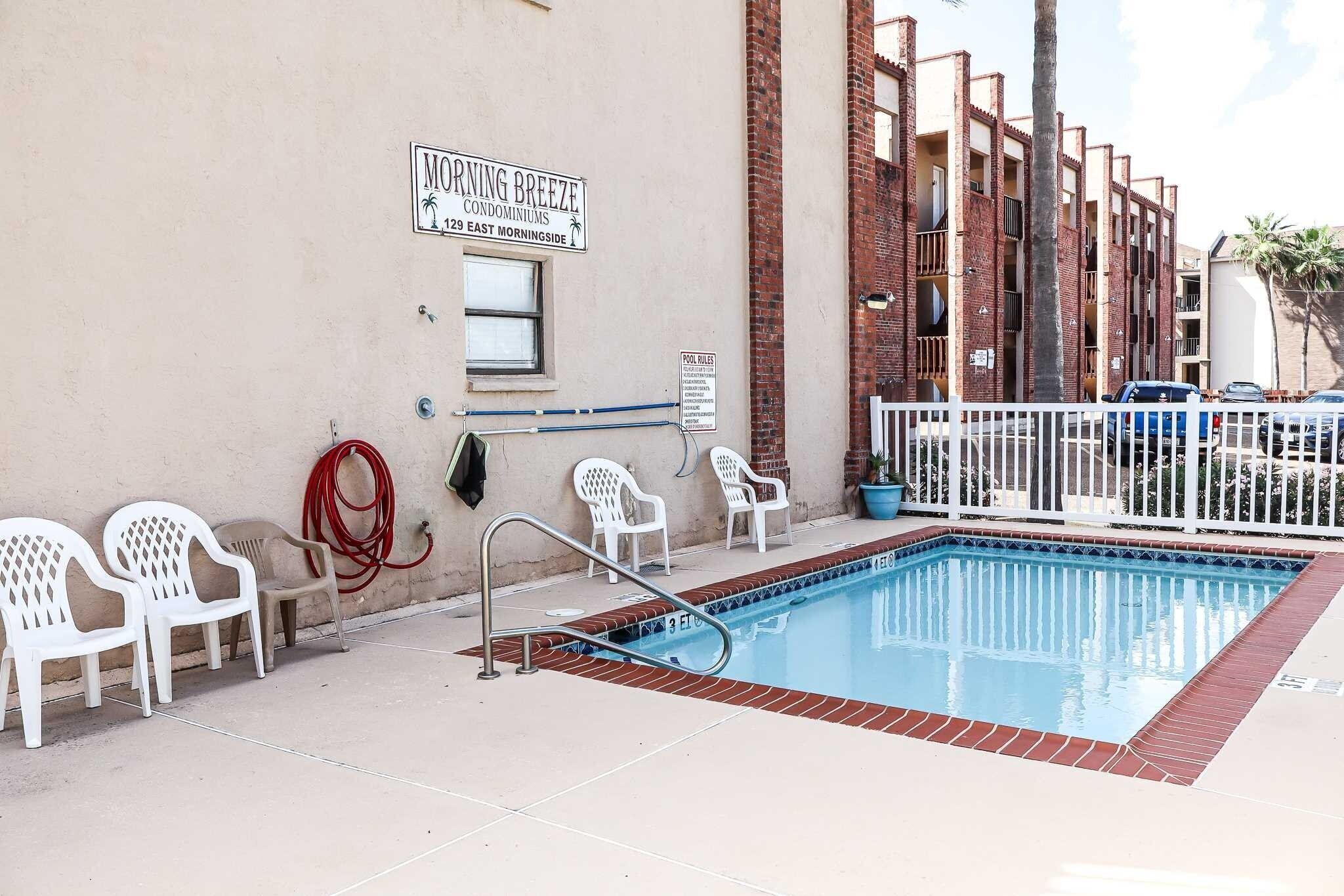 A swimming pool with a white fence, a sign for 'Morning Breeze Condominium,' and a 'Pool Rules' sign.