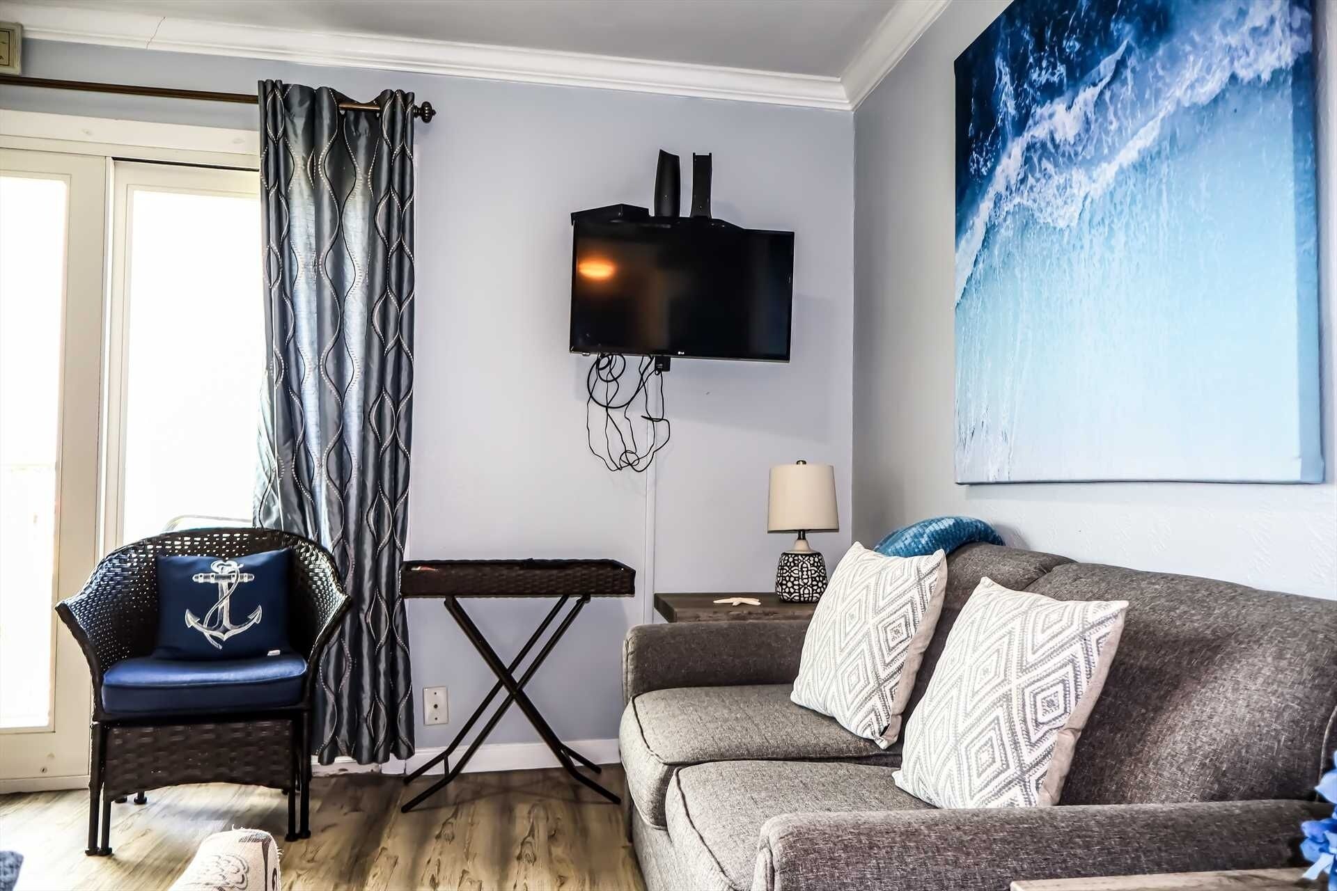 A living room with a grey sofa, a chair with an anchor design, a TV mounted on the wall, and a large wave painting.