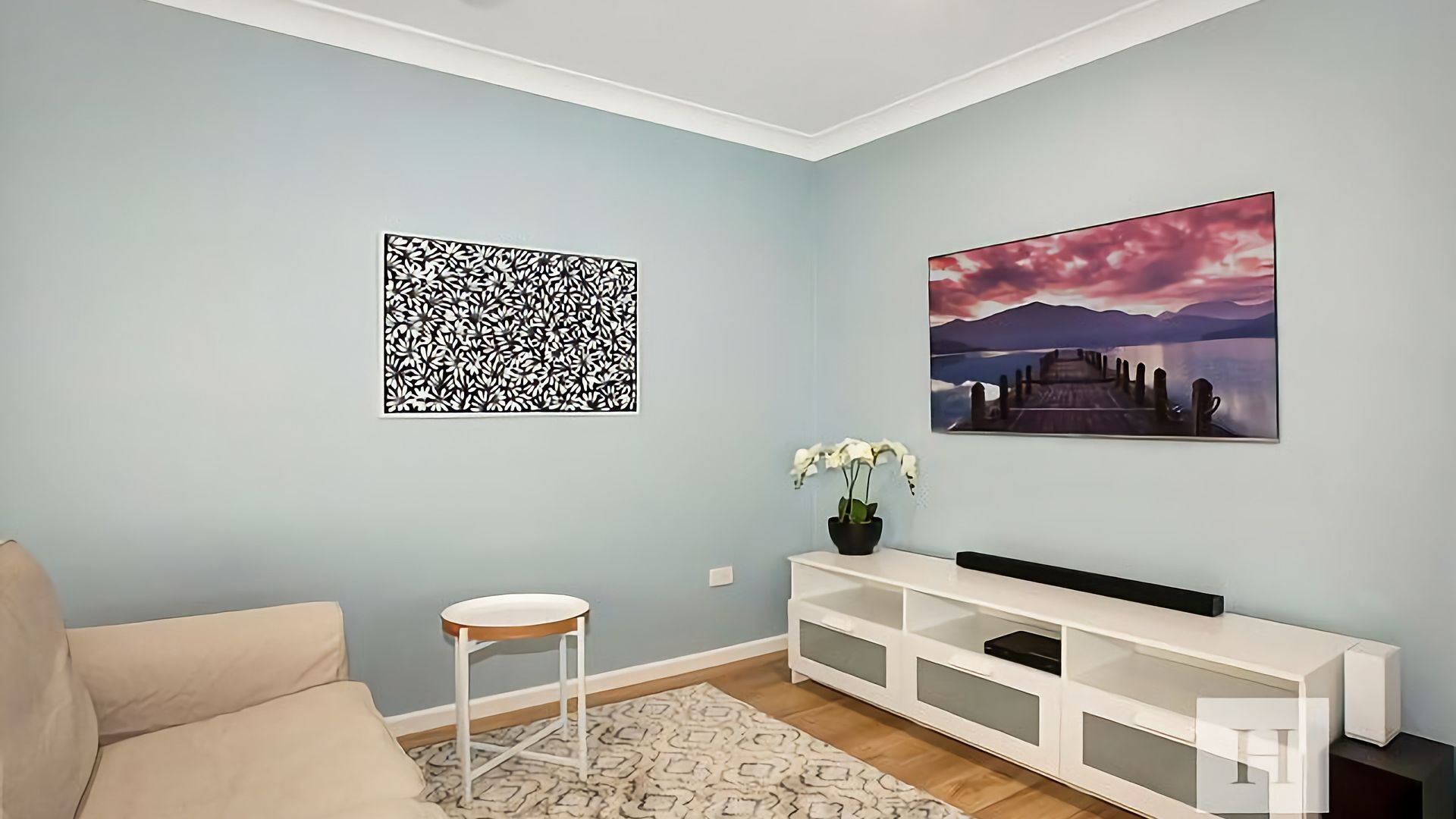 A living room with a sofa, a small white side table, a white entertainment unit, a patterned rug, and two wall art pieces.