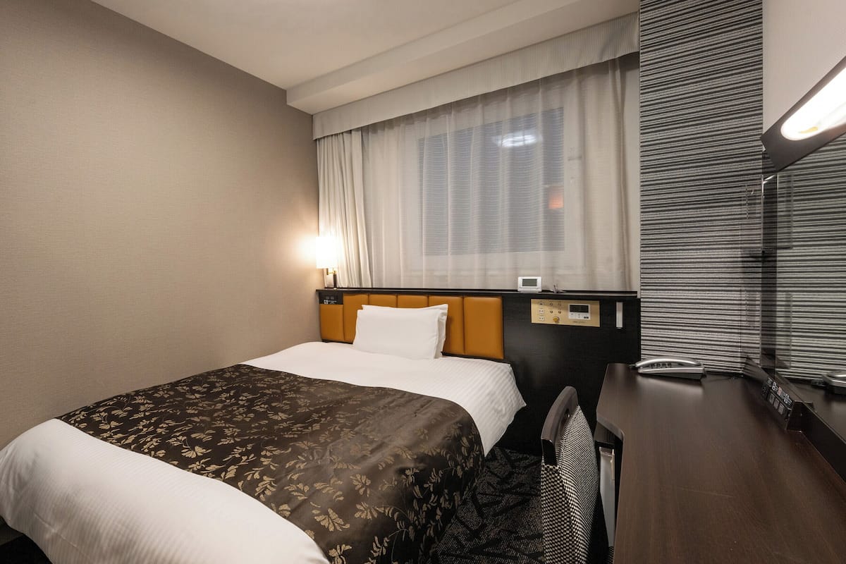 A hotel room with a bed, a desk, a chair, a window with curtains, and a wall-mounted control panel.