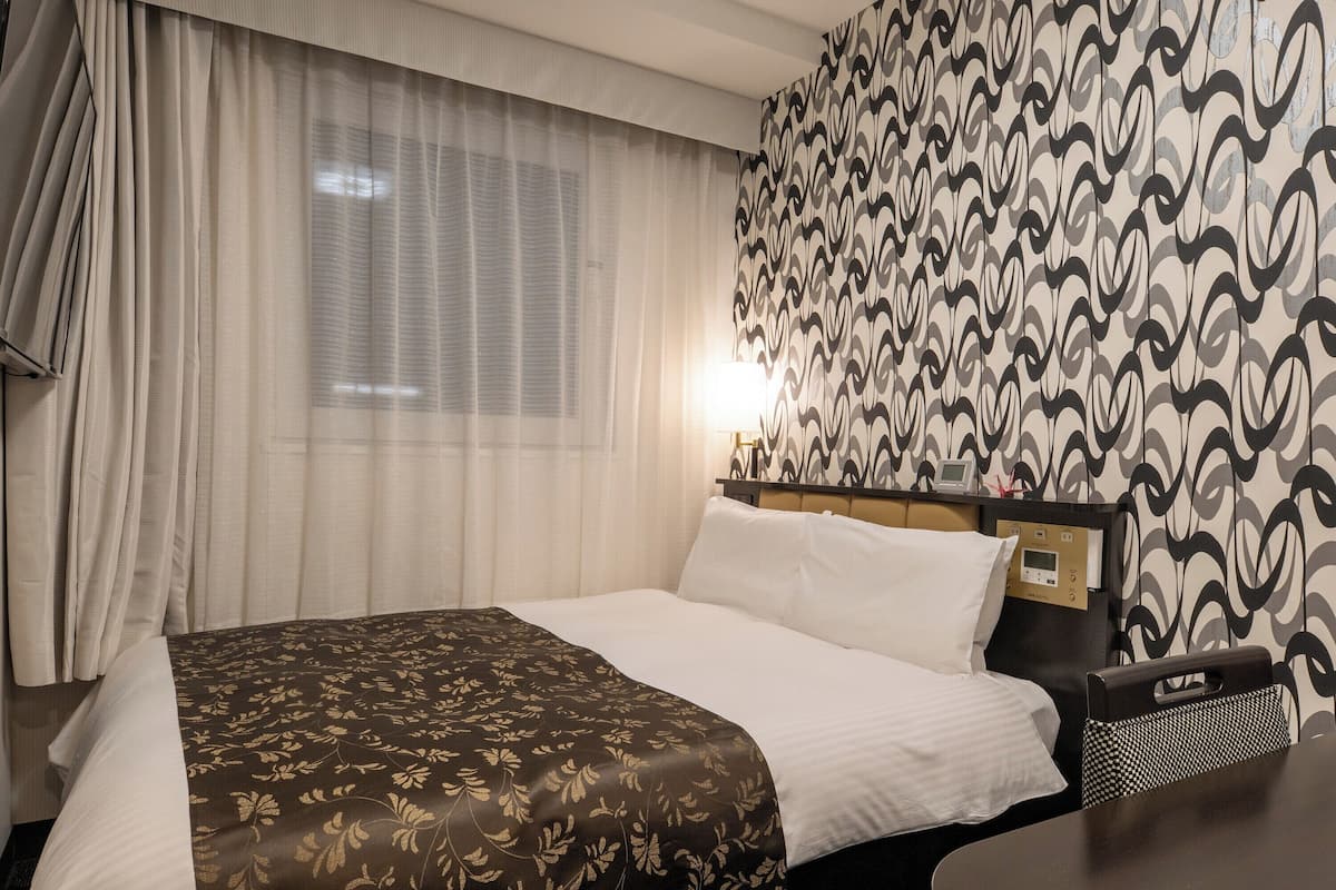 A hotel room with a bed, curtains, a patterned wall, and a bedside table with a lamp.