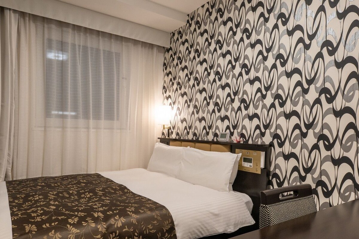 A hotel room with a bed, curtains, a patterned wall, and a bedside table with a lamp.