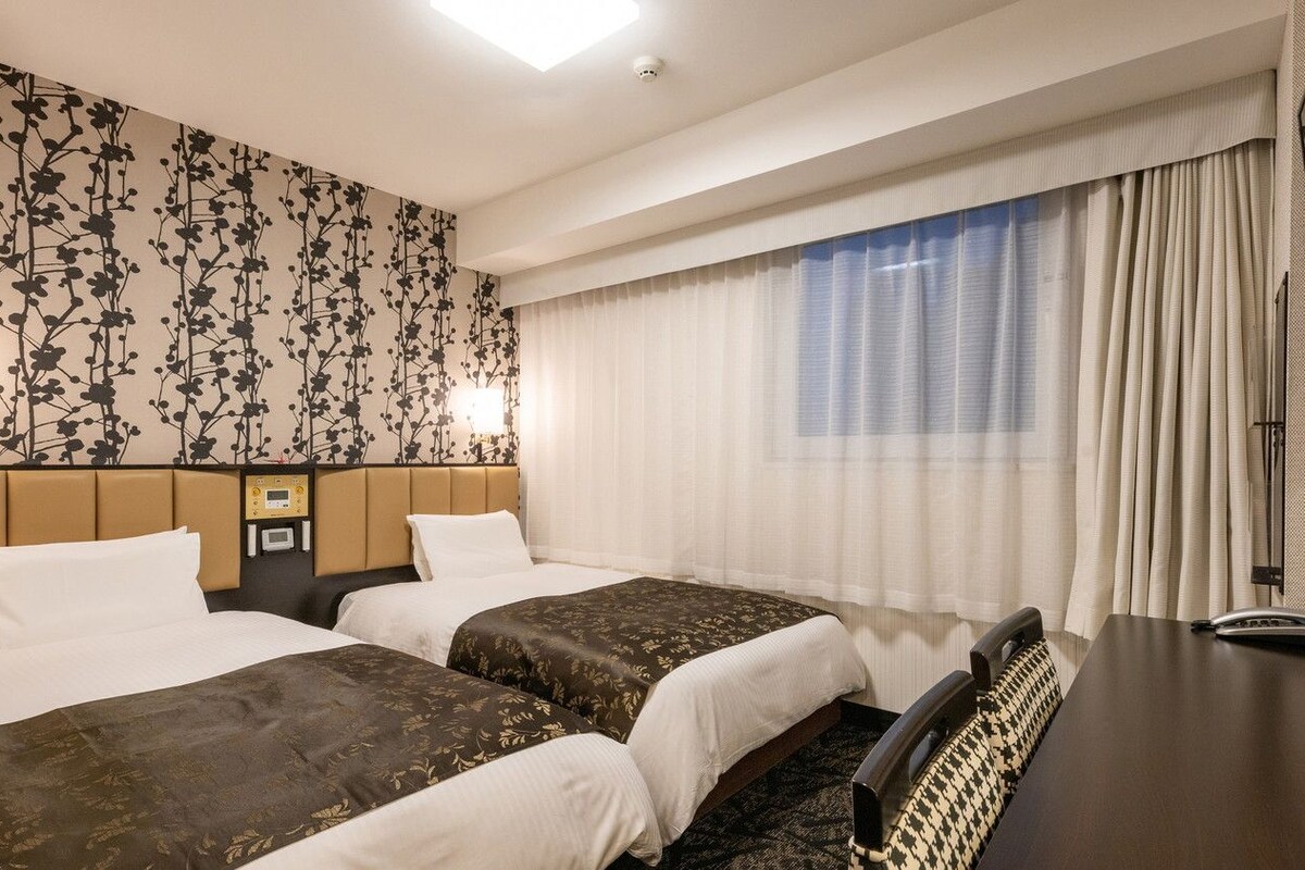 A hotel room with two beds, a desk, and a large window with curtains.