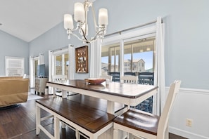 A dining area with a wooden table, white chairs, a chandelier, and a view of buildings through large windows.