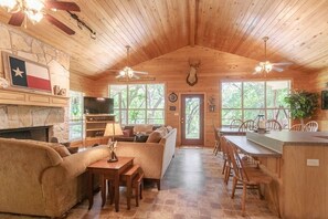 A cozy cabin interior with a stone fireplace, a sofa, a dining table, and a kitchen area with a bar. - Wagon Wheel Rr9084ww 3 Bedroom Home by RedAwning (New Braunfels)