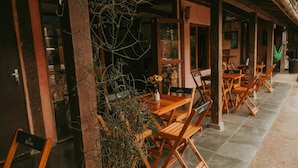 An outdoor seating area with wooden tables and chairs, some with 'Corona' branding, and a large plant in the foreground. - Drifter Hostel (São Sebastião)
